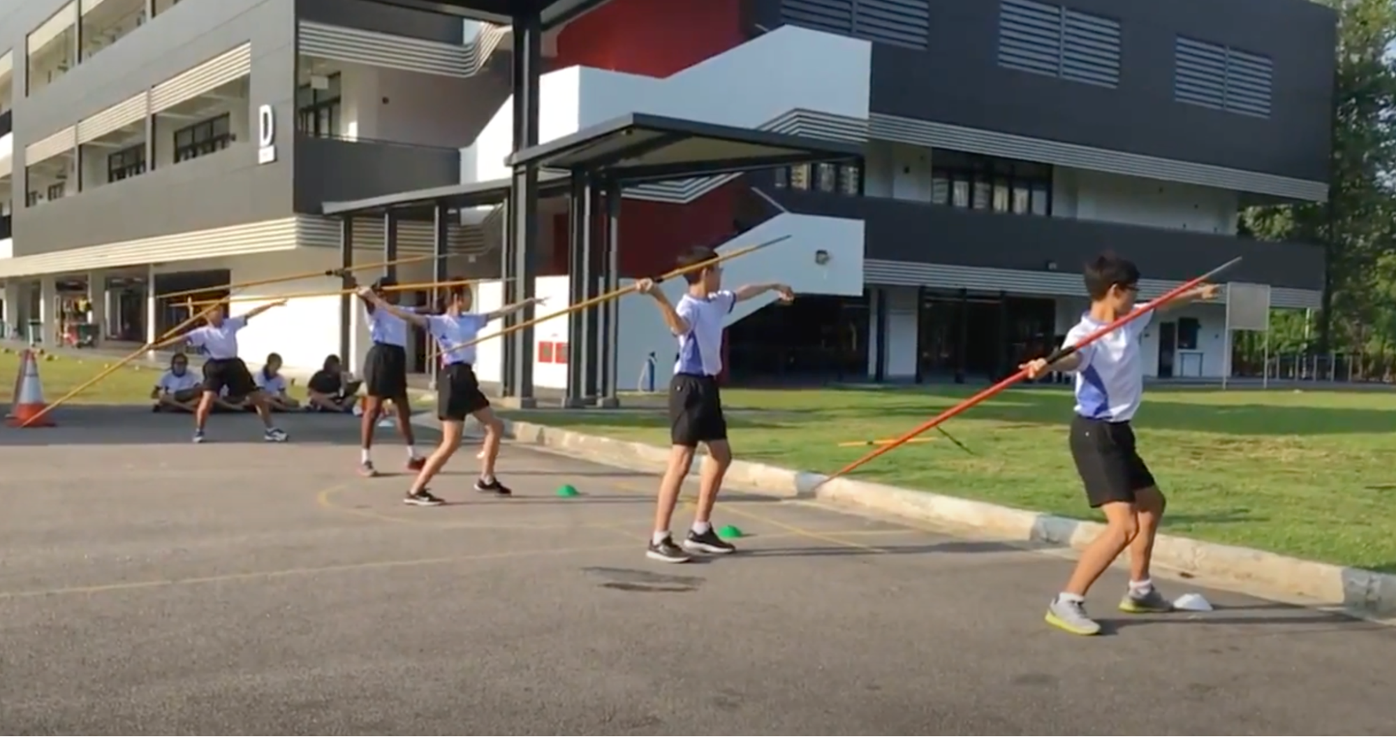 Students in uniforms practice javelin throwing stance outdoors on a school grounds with a multi-story building behind them.